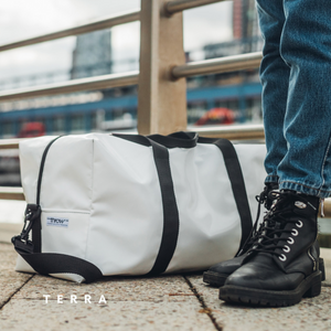 Person wearing black boots next to a white THROW duffel bag with black straps on a city street.