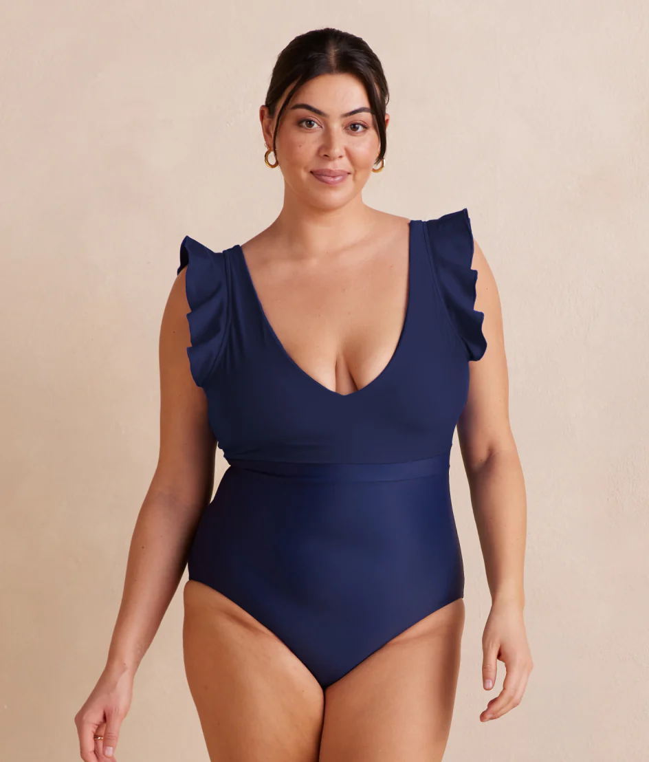 Woman wearing a summersalt navy blue swimsuit with ruffled shoulders against a beige background